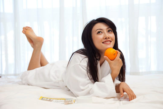 穿浴袍的年轻女人拿着橙子趴在床上