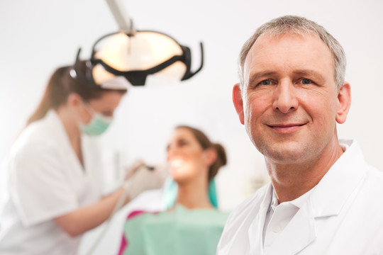 牙医在他的手术中看着观众，背景中他的助手正在给一位女病人治疗