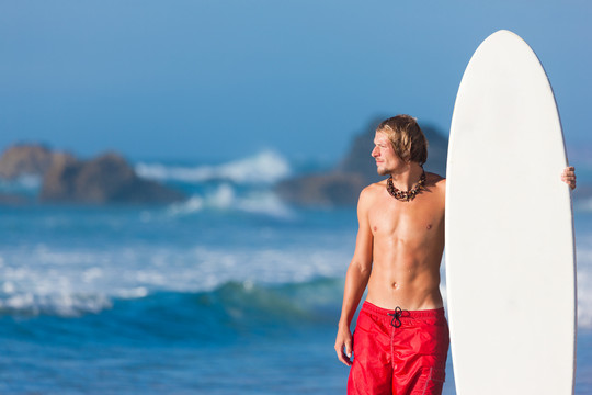 年轻的冲浪者站在沙滩上，旁边放着冲浪板，看着大海