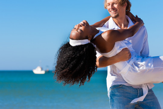 恋爱中的一对-一个白人男子背着他的非洲裔美国女人在海滩上的蓝天下