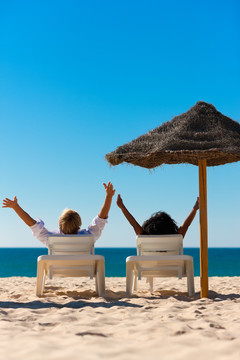 一对情侣坐在沙滩阳伞遮阳伞下的太阳椅上，舒展着双臂，感觉很自由
