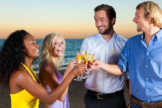 人们（两对情侣）在沙滩上开派对，在夕阳下喝酒，玩得很开心，他们穿着时髦的休闲服，喝着香槟
