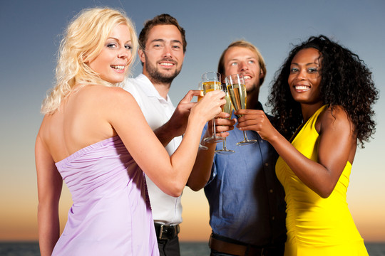 人们（两对情侣）在沙滩上开派对，在夕阳下喝酒，玩得很开心，他们穿着时髦的休闲服，喝着香槟