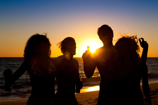 海滩上的人们（两对情侣）举行聚会，在夕阳下喝酒，玩得很开心（只能看到人们的侧影，人们手里拿着瓶子，阳