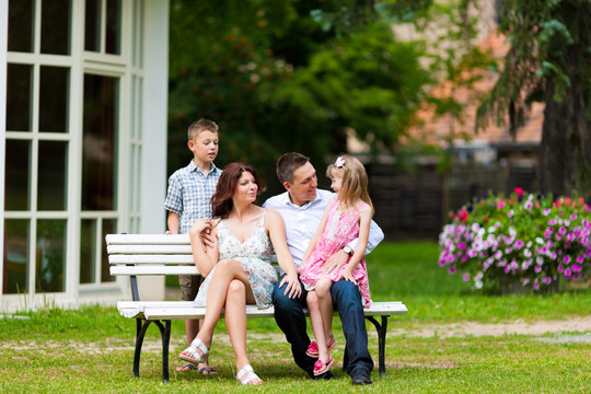 年轻幸福的一家人坐在阳光下，在新家门口的长凳上——这是一栋别墅