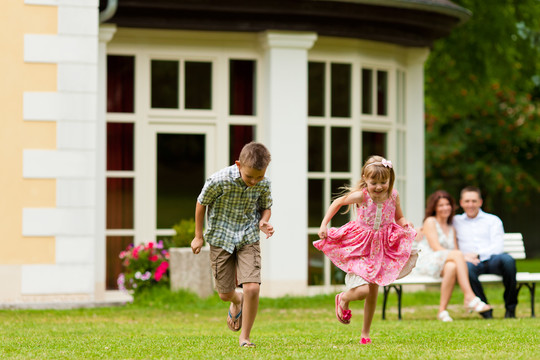 年轻快乐的家庭——孩子们在玩耍，父母坐在别墅前的长凳上晒太阳