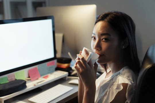 一位泰国亚裔妇女坐在办公室的电脑前，一边喝着杯子里的热咖啡。夜班时间。