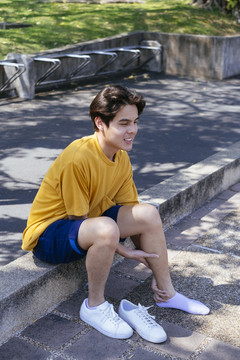 穿着黄色t恤和蓝色裤子的男孩坐在绿色公园的人行道上，在树荫下按摩脚踝。