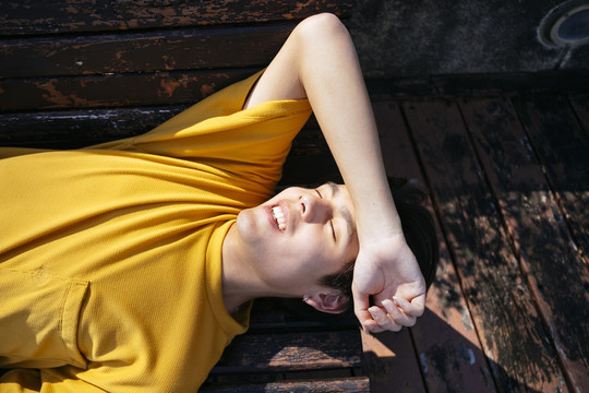 一个穿着黄色t恤和蓝色裤子的家伙在阳光下躺在木凳上，抬起手臂放在前额。