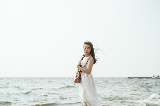 在风大的日子里，站在海边的一位身穿白裙的黑发女子抱着小提琴站着。