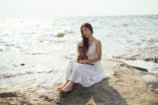 黑发白衣女子背着小提琴坐在海边的岩石上晒太阳。
