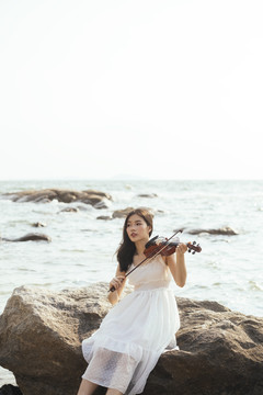黑色长发白衣女子坐在岩石上拉小提琴微笑，半海半空为背景。