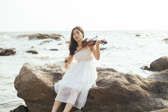 黑色长发白衣女子坐在岩石上拉小提琴微笑，半海半空为背景，横着。
