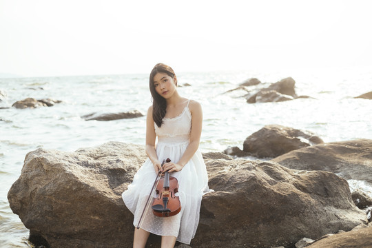 长发白衣女子从拉小提琴休息，坐在海边的岩石上，看镜头。