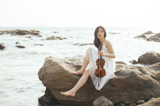 白衣美女抱着小提琴，性感地坐在岩石上。