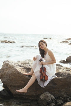 白衣美女抱着小提琴，性感地坐在岩石上。