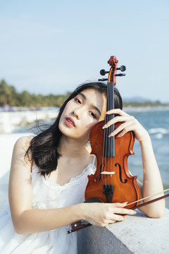一位身穿白色连衣裙的黑发女子坐在大理石长凳上，在阳光下把头靠在小提琴上。