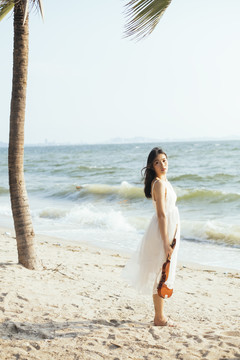 身穿白裙的黑长发女子手持小提琴站在沙滩上看着镜头。
