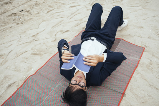 穿深色西装的黑发男人躺在垫子上，在沙滩上看书。