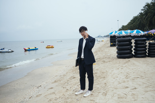 一个穿着深色西装的黑发男人站在沙滩上，在模糊的天空下和他的顾客通电话。