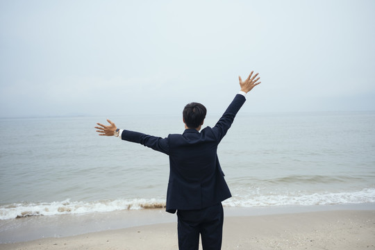 一个黑发的穿着深色西装的家伙站在沙滩上，张开双臂捂着头，像是在欢迎海上的风，水平的。