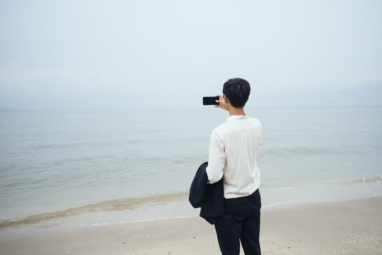 黑发白衣人站在阴沉的沙滩上，用智能手机拍下大海的照片。
