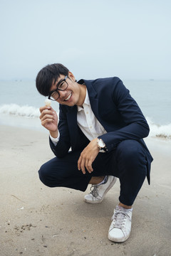 黑发男身穿深色西装坐在沙滩上，面带微笑地展示手中的白色贝壳。