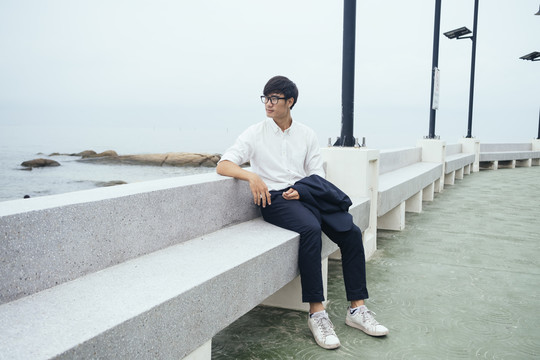 黑发汉子穿着白衬衫坐在大理石长凳上沿着码头在大海中布满了岩石。
