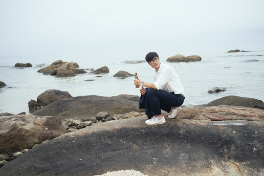 穿着白衬衫的黑发男人坐在海边的岩石上，手里拿着瓶子。