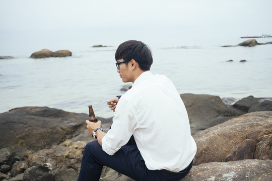黑发小伙穿着白衬衫坐在海边的岩石上，手里拿着酒瓶，望着大海，横。