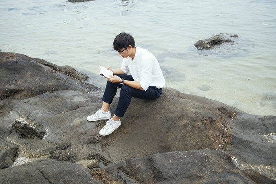 穿着白衬衫的黑发男人坐在海边的岩石上看书。