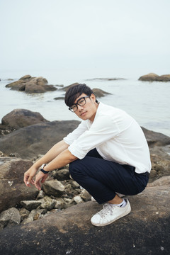 黑发人穿着白衬衫，坐在海边的岩石上，双臂放在膝盖上。