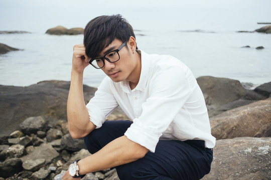 穿着白衬衫的黑发男人坐在海边的岩石上想着他的工作。