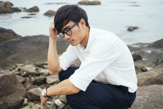 穿着白衬衫的黑发男人坐在海边的岩石上想着他的工作。