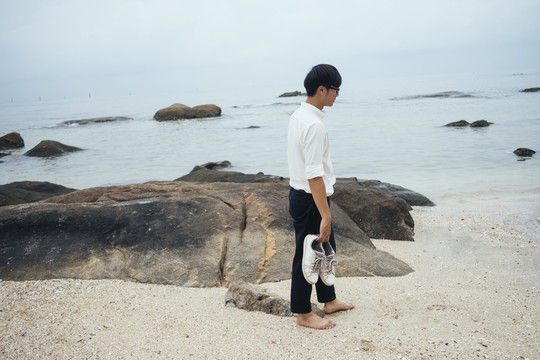 穿白衬衫的黑发男人脱下鞋子，不穿鞋子走在海边有岩石的海滩上。