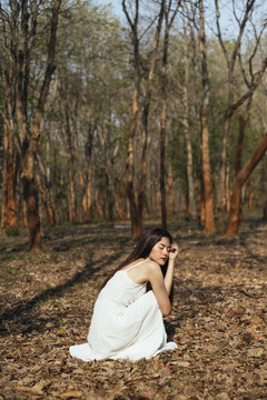 亚洲美丽的年轻女子穿着优雅的白裙坐在秋林里。在森林里迷路的女孩。
