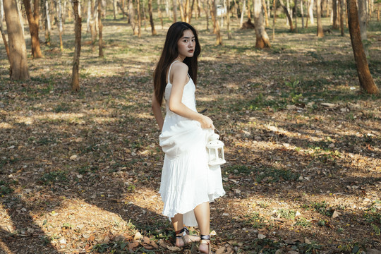 美丽的年轻女子穿着优雅的白裙，提着白灯笼走在森林里。