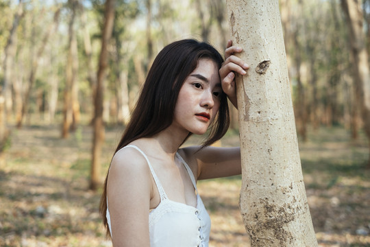 美丽的年轻女子穿着优雅的白色连衣裙，倚在树上，神情忧虑。