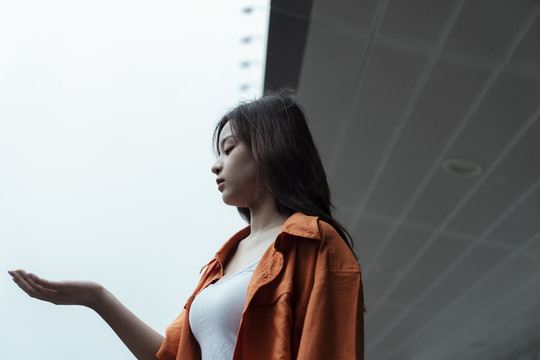 橘色衬衫女孩站在商场的雨篷前，伸出手查看雨水。