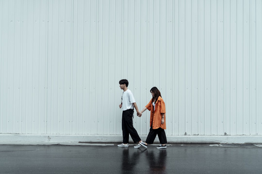 一对年轻的夫妇沿着白墙走在一起，手牵着手。