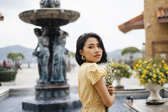 年轻的亚洲女人穿着黄色的裙子，手持白色的花束站在室外的喷泉旁。