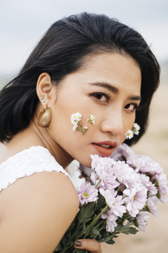 穿着白衬衫和蓝色牛仔裤的年轻性感亚洲女子的肖像，手持一束鲜花，倚在户外的白色栅栏上。
