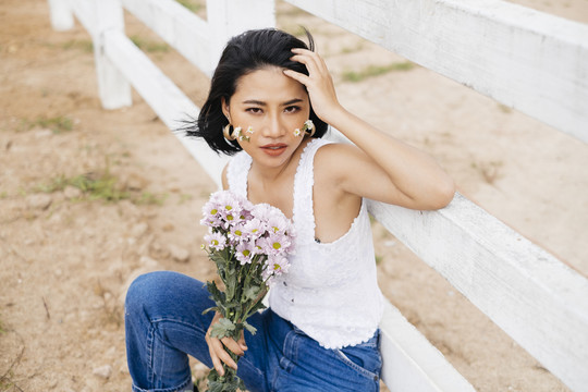 穿着白衬衫和蓝色牛仔裤的年轻性感的亚洲女人坐在白色的篱笆上，手里拿着一束花。