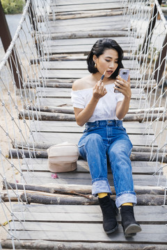 性感的亚洲女人坐在木桥上，整容，从旁边的小篮子里拿起设备。