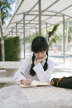 扎着辫子、留着流苏头发的女学生坐在公园的大理石长凳上，一边做作业，一边等着她的父母。