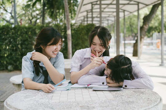 三个穿着不同制服的漂亮亚泰学生放学后一起做作业。书呆子女孩在桌子上睡着了。另外两个在恶作剧。
