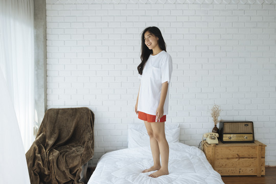 穿着睡衣的可爱亚洲女人站在卧室舒适的床上微笑。