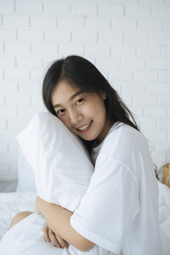 可爱的亚洲小姐抱着白色的枕头，在卧室里微笑。