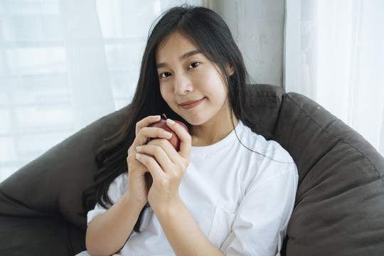 可爱的亚洲年轻女子双手捧着苹果坐在舒适的沙发长凳上。