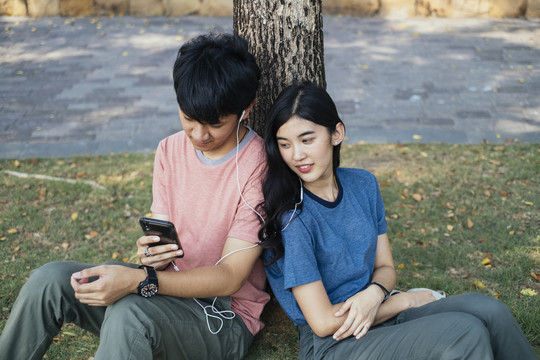 一对亚泰夫妇坐在树下，一起用耳机听音乐。浪漫时刻。